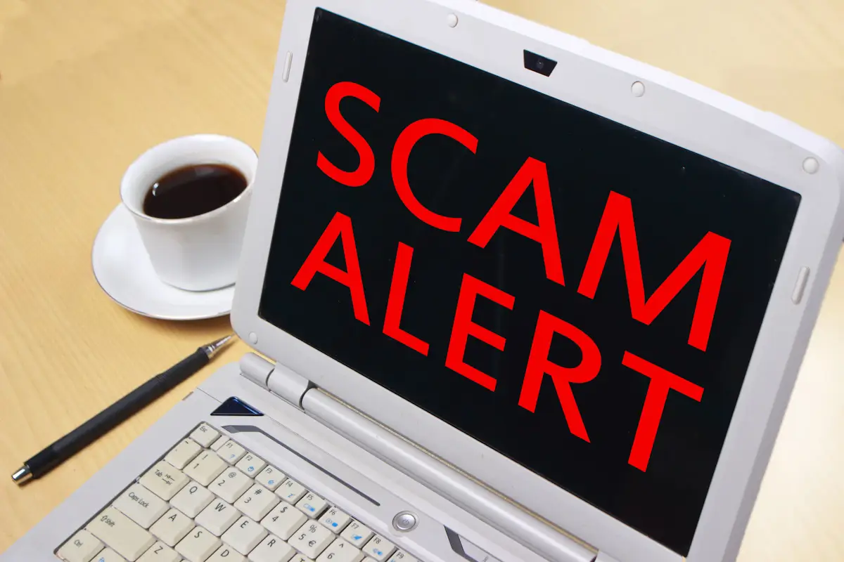 Laptop screen displaying "SCAM ALERT" with coffee cup.