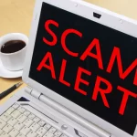 Laptop screen displaying "SCAM ALERT" with coffee cup.